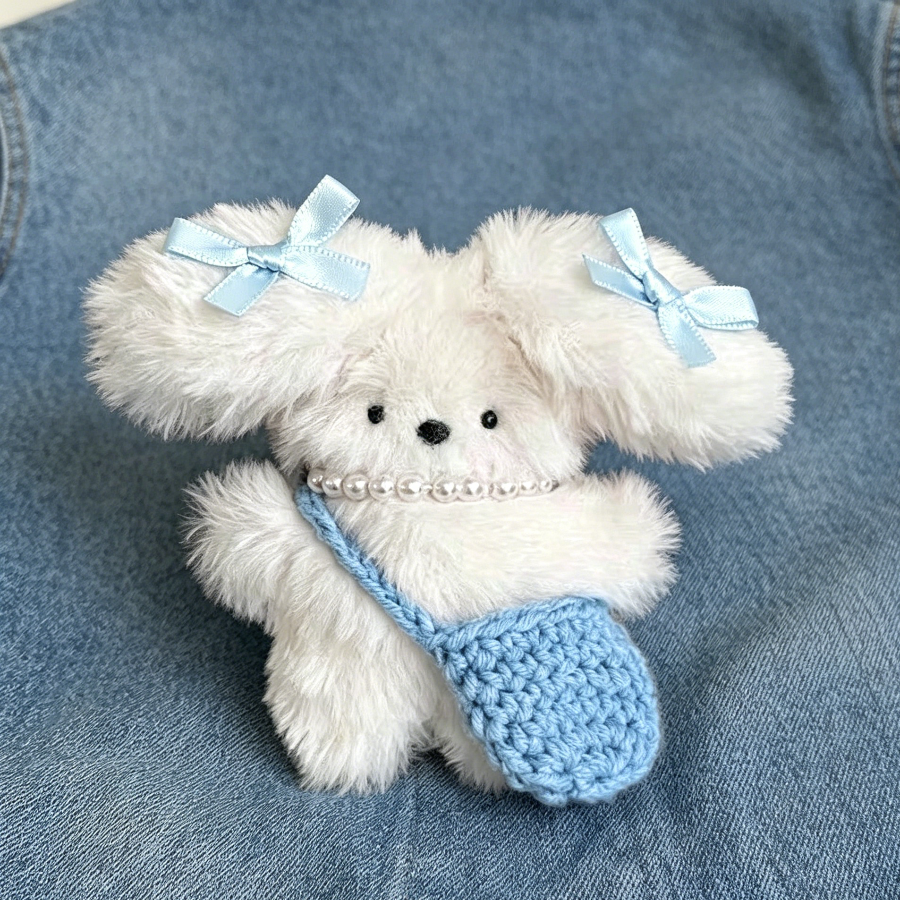 Cream plush bag charm with blue bows, pearl necklace, and a tiny crocheted blue bag attached to a metal clasp.