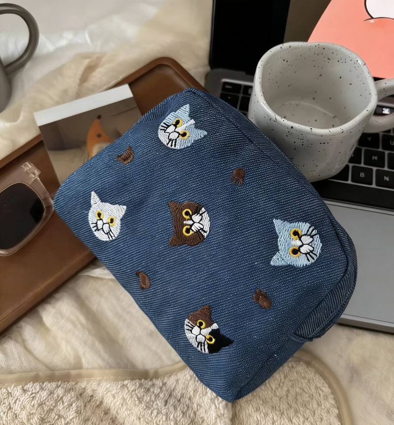 Three pouches in white, denim blue, and lavender, each with embroidered cat faces and paw prints