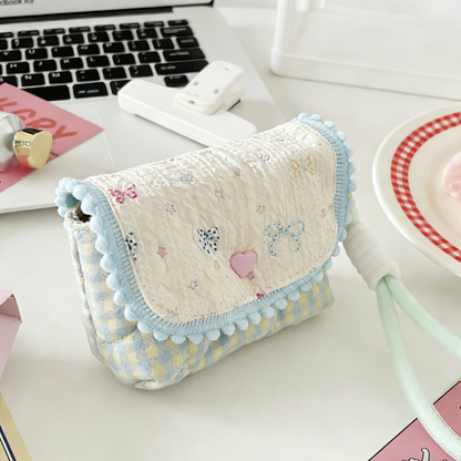 Four small quilted pouches in pastel colours, each with a patterned flap, pom‑pom trim, heart button, and matching wrist strap