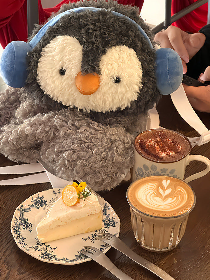 Fluffy grey penguin mini backpack with blue earmuffs worn over the shoulder with white straps