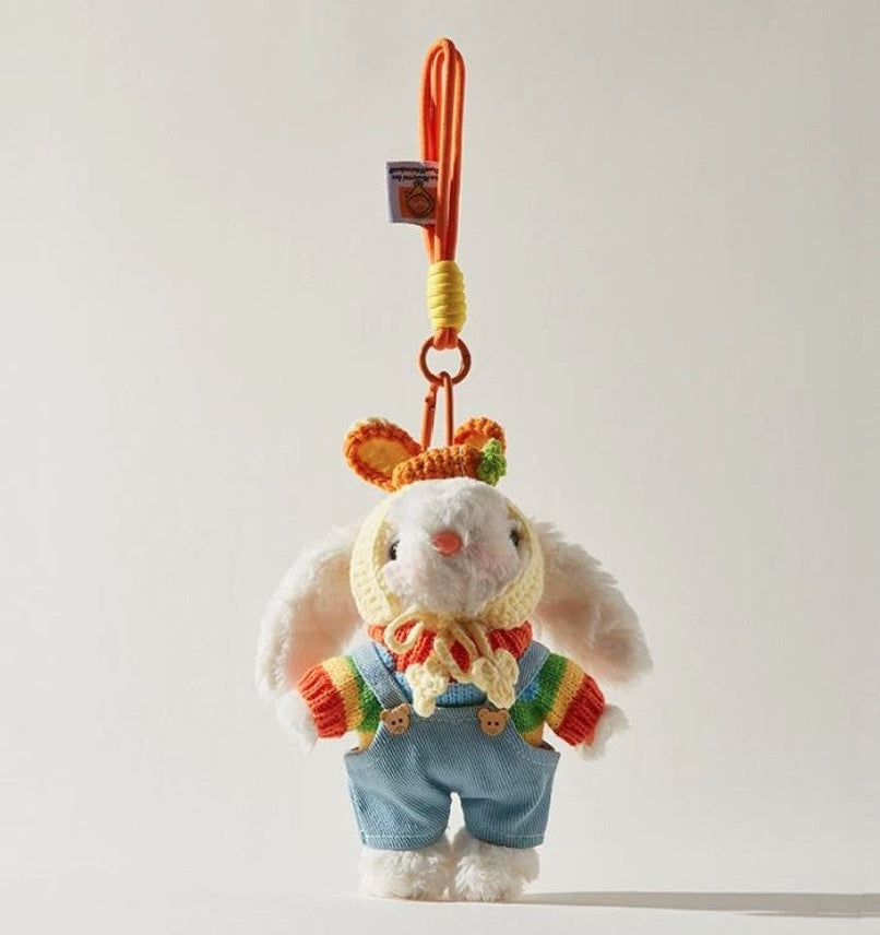 White plush bunny bag charm wearing a rainbow sweater, blue overalls, and a yellow crocheted bonnet, attached to an orange strap