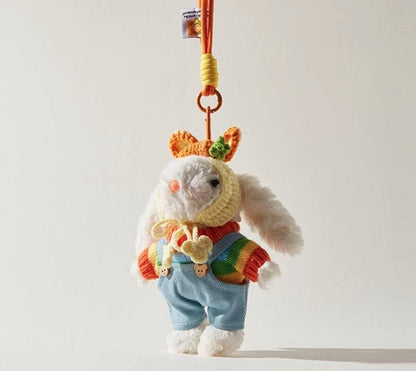 White plush bunny bag charm wearing a rainbow sweater, blue overalls, and a yellow crocheted bonnet, attached to an orange strap