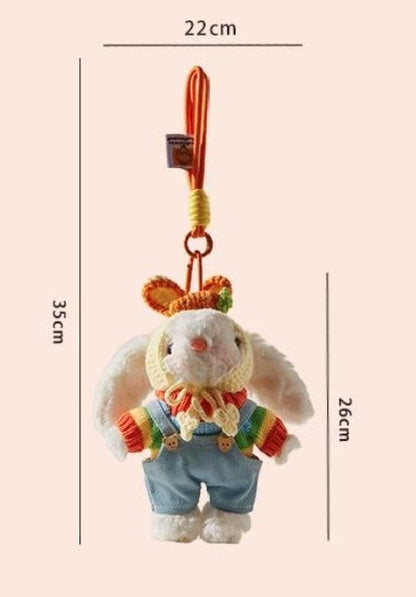 White plush bunny bag charm wearing a rainbow sweater, blue overalls, and a yellow crocheted bonnet, attached to an orange strap