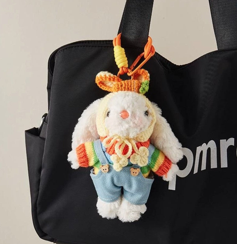 White plush bunny bag charm wearing a rainbow sweater, blue overalls, and a yellow crocheted bonnet, attached to an orange strap