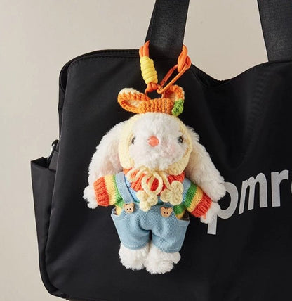 White plush bunny bag charm wearing a rainbow sweater, blue overalls, and a yellow crocheted bonnet, attached to an orange strap