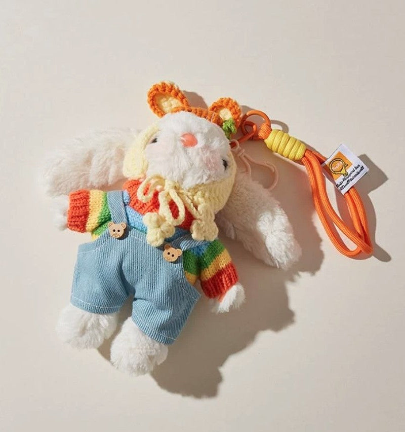 White plush bunny bag charm wearing a rainbow sweater, blue overalls, and a yellow crocheted bonnet, attached to an orange strap